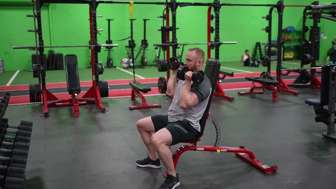 Person performing a seated Arnold press on an upright bench, holding dumbbells in front of their shoulders with palms facing inward and elbows bent, preparing to rotate their arms and press the weights overhead.