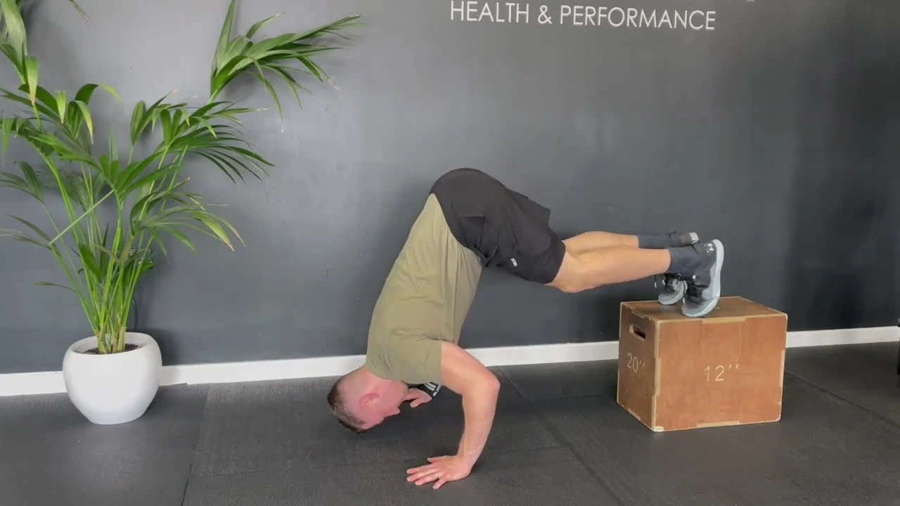 Person performing an elevated pike push-up with feet on a wooden box, hips raised and head lowered toward the floor, demonstrating the shoulder-focused exercise.