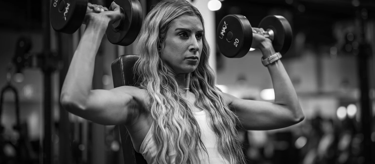 Woman performing a seated dumbbell shoulder press in a gym, holding weights at shoulder height.