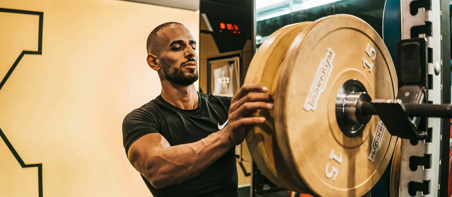 A man in a gym sliding a fifteen kilo plate onto the end of a loaded barbell resting in a squat rack.