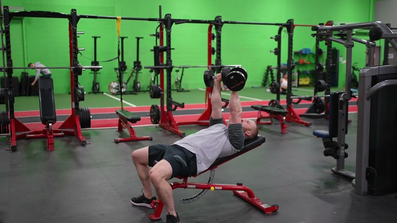 Person performing an incline dumbbell bench press while lying on an inclined weight bench, holding two dumbbells directly above their upper chest with arms extended and feet planted on the floor.