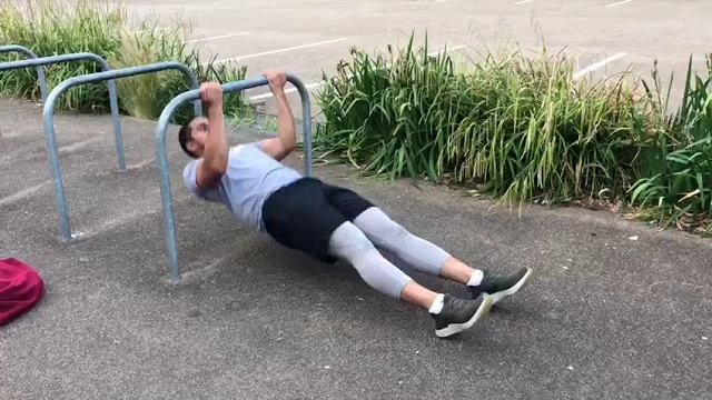 Person performing an inverted bicep curl using an outdoor horizontal bar, pulling their forehead toward the bar with an underhand grip while keeping their body straight, legs extended, and heels on the ground.