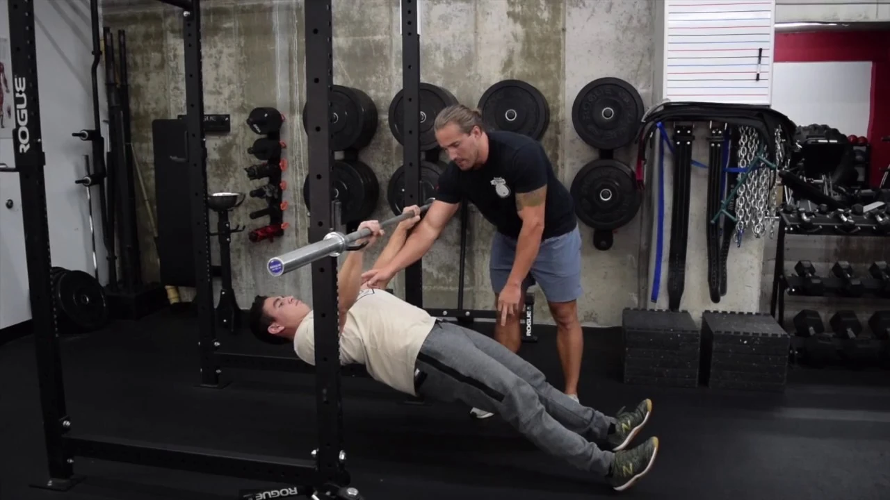 Person performing an inverted row using a barbell secured in a squat rack, pulling their chest toward the bar while keeping their body straight and heels on the floor, with a trainer observing their form.