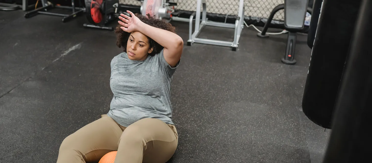 Woman sitting on the gym floor with her arm resting across her forehead after a tiring workout.
