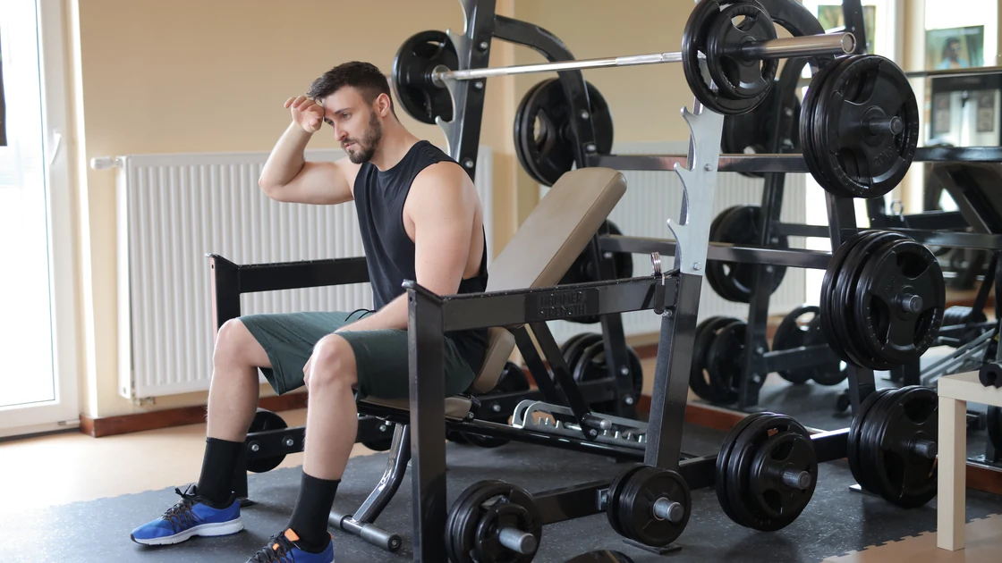 Man sitting on a gym bench with his hand against his forehead, resting beside a loaded barbell.