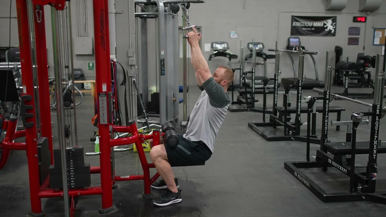 Person performing a seated neutral-grip lat pulldown on a cable machine, holding the handles with palms facing inward and arms extended overhead, demonstrating the starting position of the exercise.