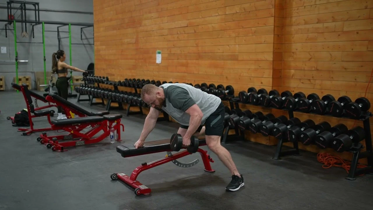 Person performing a single-arm dumbbell row with one hand supported on a flat bench, pulling a dumbbell toward their hip while keeping their back straight and feet planted on the floor.