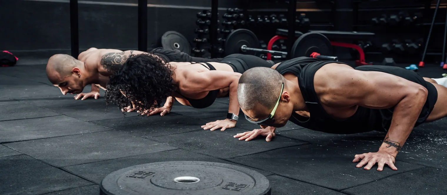 Three people performing push-ups on the floor of a gym, lowering their chests toward the ground.