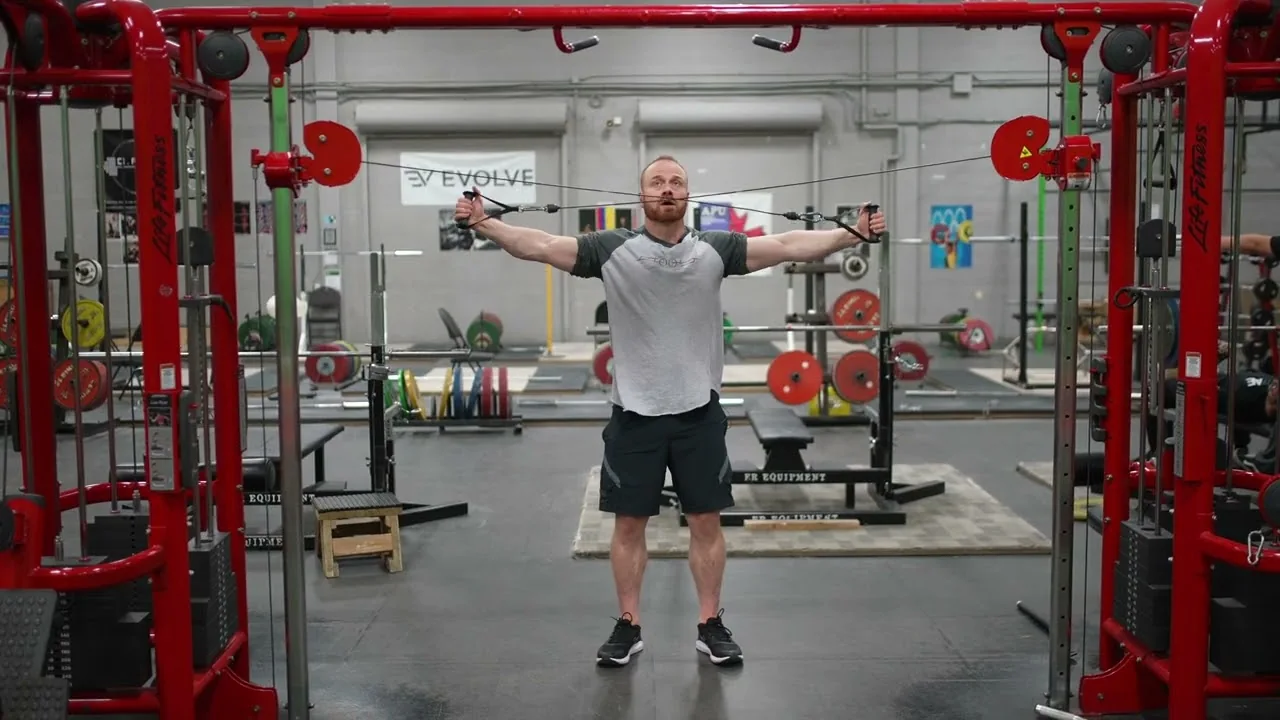 Person performing a standing reverse cable crossover on a dual cable machine, holding the handles with arms extended out to the sides at shoulder height, targeting the rear deltoids and upper back.