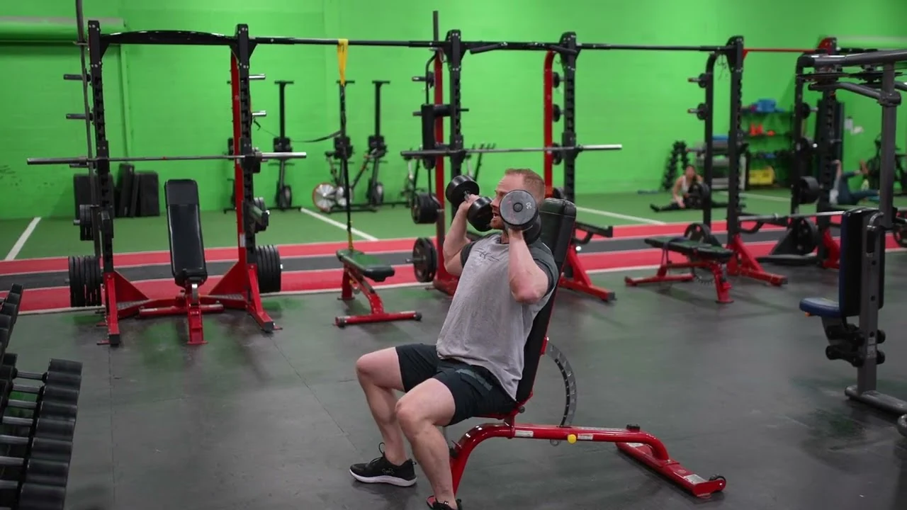 Person performing a seated dumbbell shoulder press on an upright bench, holding two dumbbells at shoulder height with elbows bent and feet planted on the floor.