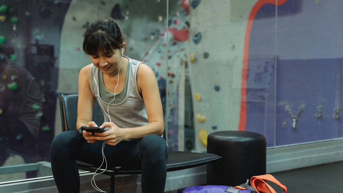 Woman sitting on a gym bench checking her smartphone during a workout, with earphones in.