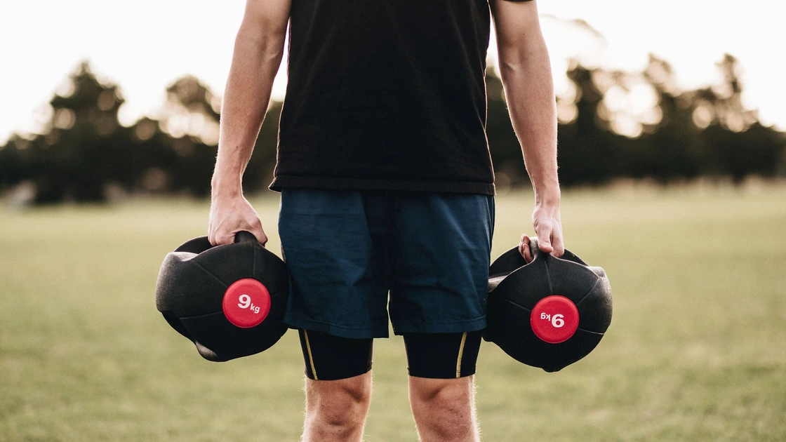 Person holding a dumbbell in each hand in an outdoor setting, demonstrating the starting position of a farmer’s carry.